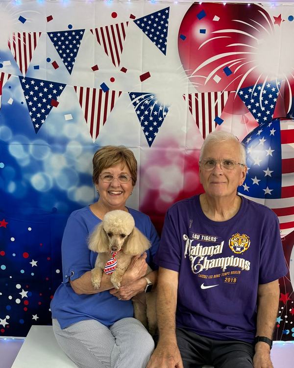 Two people sit with a small cream toy poodle wearing a patriotic necktie by a July 4th backdrop.