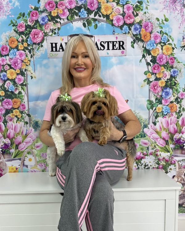A woman holds two Yorkshire terriers wearing green bows against a floral Happy Easter backdrop.