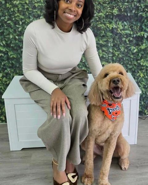 A person sitting beside a fluffy apricot goldendoodle in front of a leafy wall and neon salon sign.