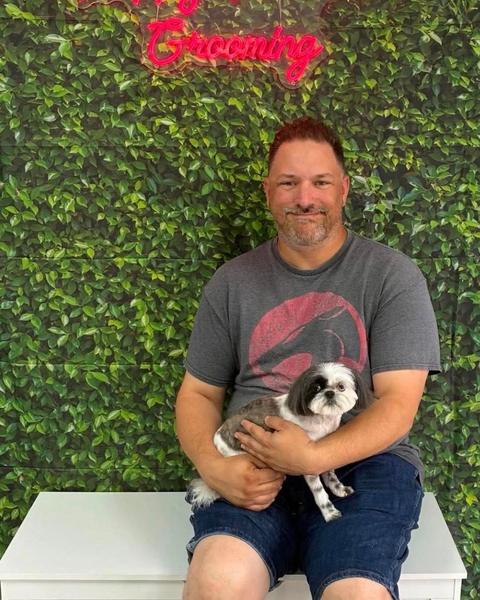 A person holding a grey and white shih tzu in front of a leafy wall and neon sign.