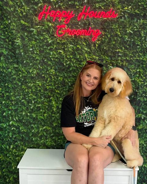A customer poses with a large cream goldendoodle against a green leaf wall and neon sign.