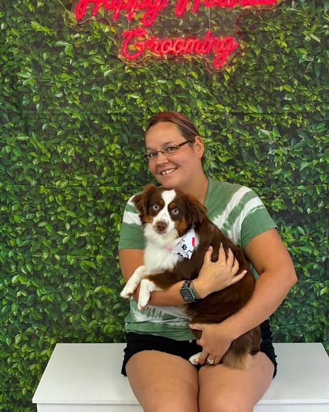 A woman holds a brown and white puppy wearing a bandana against a green leaf wall.