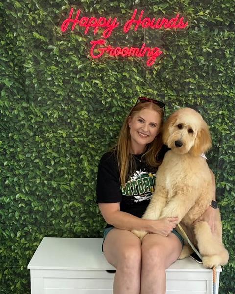 A woman sits with a cream goldendoodle with a fluffy teddy bear cut against a green leaf wall.