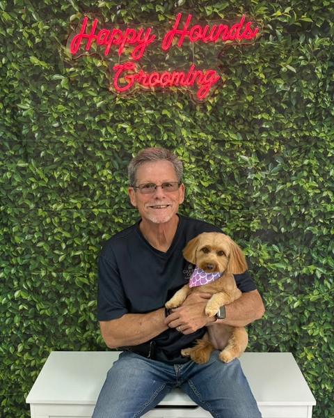 A man holds a small tan and cream dog wearing a purple bandana against a green leaf wall.