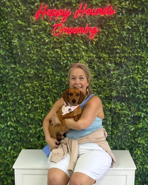 A woman holds a small red dachshund puppy wearing a bandana against a green leaf wall.