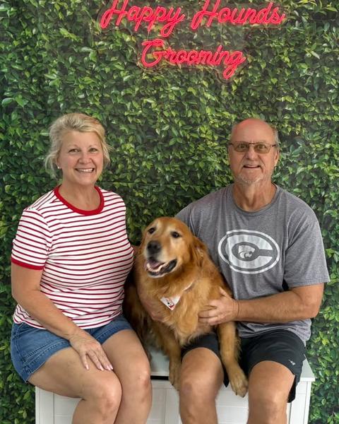 A man and woman sit with a golden retriever between them against a green leaf wall.
