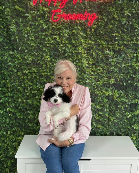 A woman holds a black and white puppy wearing a bow tie against a leaf wall.