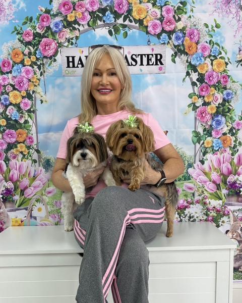 A woman holds two Yorkshire terriers wearing green bows against a floral Happy Easter backdrop.
