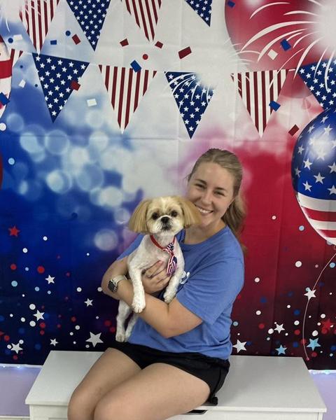 A customer holds a freshly groomed cream dog with a teddy bear trim against a patriotic backdrop.