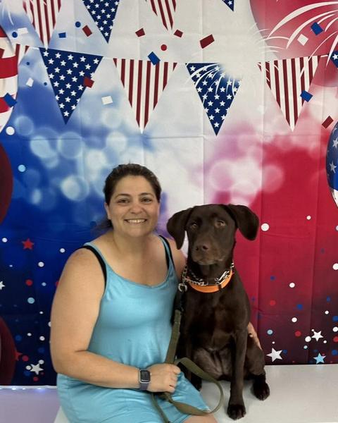 A customer smiles beside a chocolate brown Labrador mix wearing an orange collar against a patriotic backdrop.