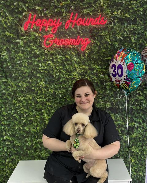 A person holds a small cream toy poodle wearing a necktie by a leafy wall and sign.