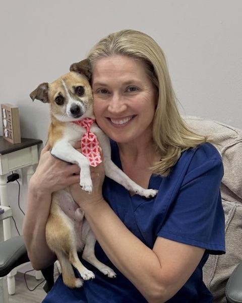 A person in scrubs holds a small tan and white dog cheek to cheek indoors.